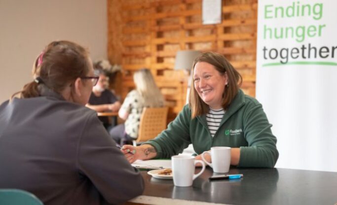 Woman wearing green fleece with Trussell Trust logo taking notes as they are sat opposite another lady with a cup of tea.