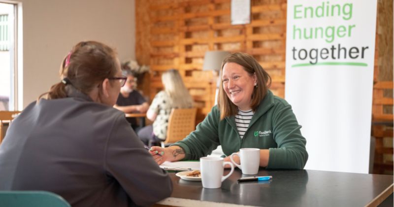 Woman wearing green fleece with Trussell Trust logo taking notes as they are sat opposite another lady with a cup of tea.
